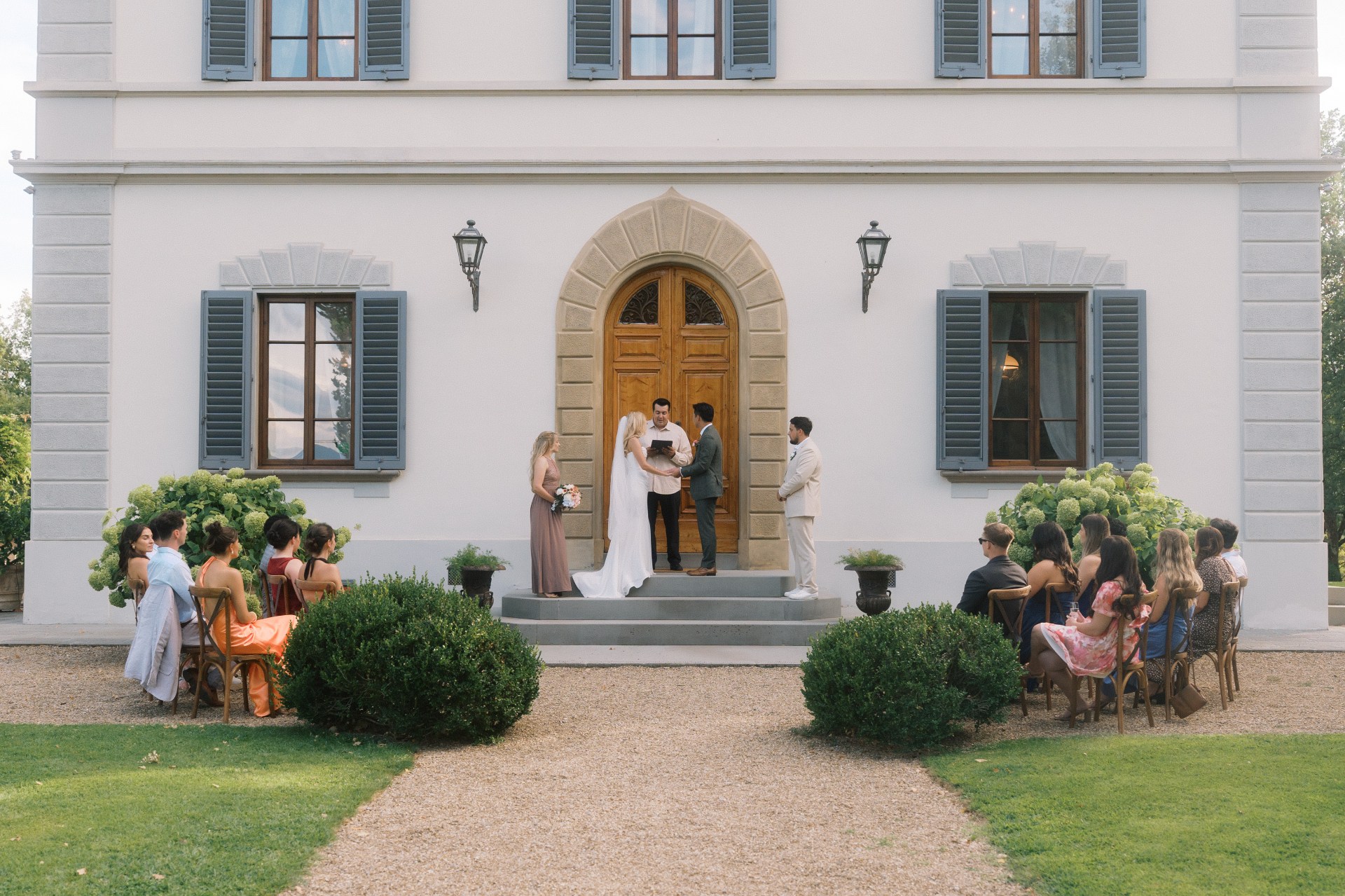 Alec Russo officiating an outdoor wedding ceremony in front of a villa with guests seated nearby